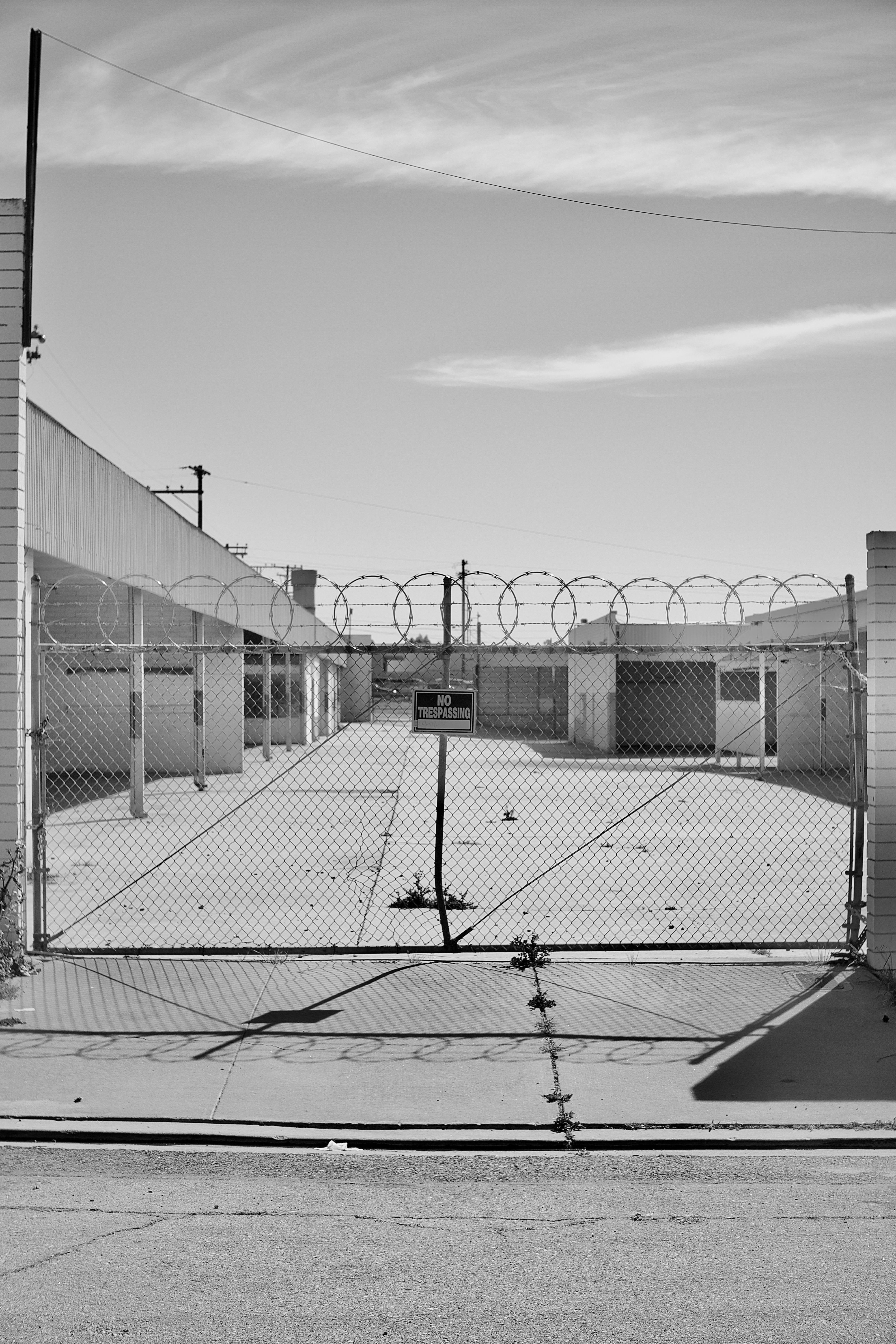 The edge of an industrial district at twilight, with a chain-link fence and weeds pushing through concrete.